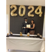 graduate posing behind table under the 2024 balloons with cap and gown on holding certificate with housing staff